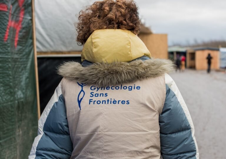 Bénévole de Gynécologie Sans Frontières vue de dos, portant le gilet de l’association dans un camp de personnes exilées